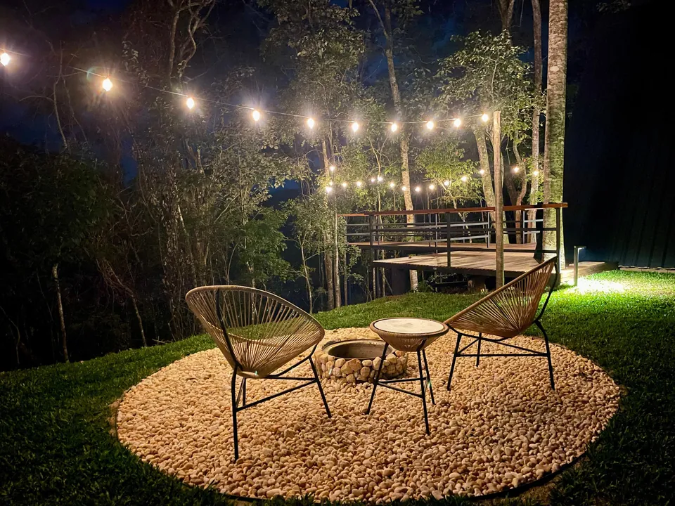 Área do fogo de chão com poltronas sob o varal de luzes, na mata, à noite