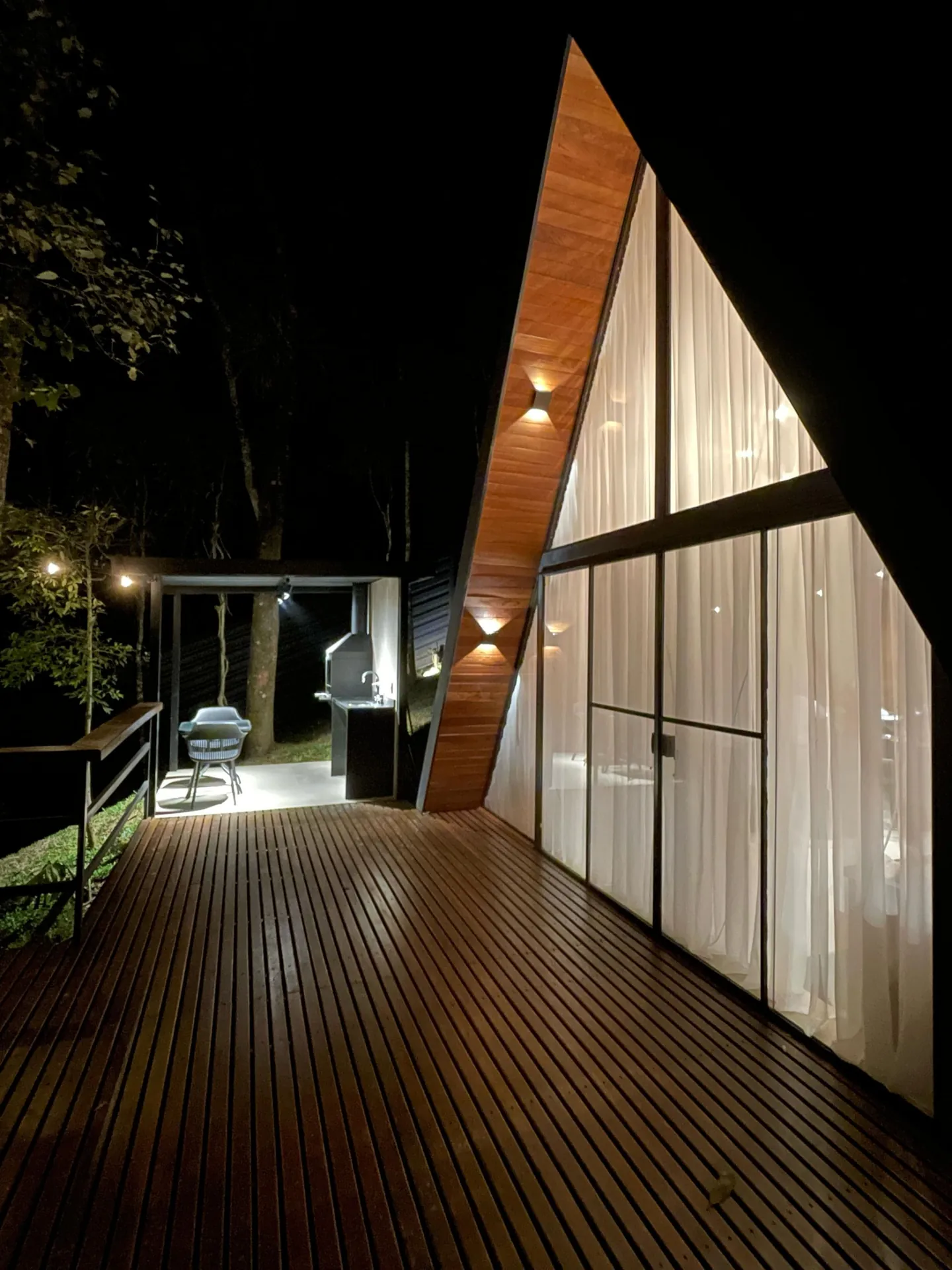 Deck de madeira iluminado à noite diante da fachada A-frame de vidro
